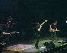 Four people. On the far left a man with a low hand. Mounted on a platform a man sits on a bench with a keyboard in front. In the center a man standing, wearing black with his left hand and taking a microphone with his right hand extended. On the far right a man semi-inclined holding a guitar.