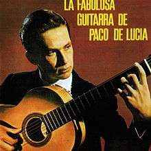 Man in black suit holding flamenco guitar in front of a reddish brown background