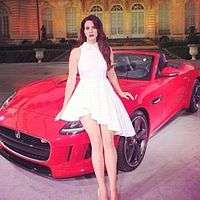 A brunette female wearing a white dress poses on a red luxury car