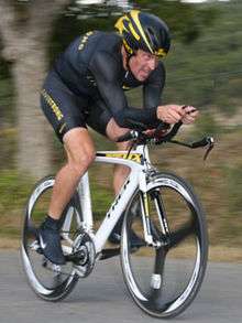 A cyclist in a black jersey with gold trim crouched into an aerodynamic position on his bicycle, riding down a road