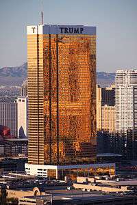 A tall rectangular-shaped tower in Las Vegas with exterior windows shimmering with 24-carat gold. It is a sunny day and the building is higher than many of the surrounding buildings, which are also towers. There are mountains in the background. This tower is called the Trump Hotel Las Vegas.
