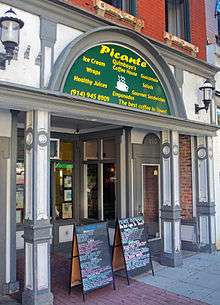 An ornate storefront with a semicircular green entablature over the entrance that says "Picante" in large letters and "Quimbaya's Coffee House" in smaller letters below it, surrounded by the names of some foods and drinks