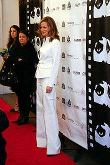 a dirty blonde woman in a white pant suit stands on a red carpet with two women in the background