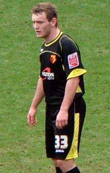 A man wearing black shirt and shorts, with yellow collar and trim, standing on a grass field