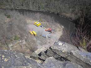 View from above of a curving river in a rocky gorge, several school buses and trucks towing trailers with boats and rafts are parked on a gravel area at left