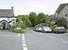 Street scene showing a road unction with parked cars. On the left and right are stone houses with trees and fields in the distance.