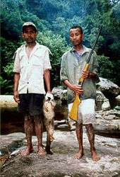 Two Malagasy hunters stand near a stream, one holding a gun, the other holding a lemur with a white head.