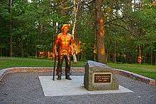 Life-size bronze statue of a shirtless man with a hat, resting his right hand on a pick axe and holding a shirt in his left hand. The top half of the statue is lit orange by the setting sun. A boulder to the right has a plaque that reads "Tioga County 'CCC Worker' 1933 - 1942".