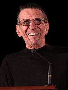 A coloured photograph of a man wearing a black jumper and glasses. He is smiling and standing on a platform.