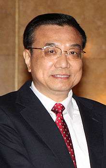 a smiling man with a short haircut, wearing glasses, a white shirt, a suit and a red tie