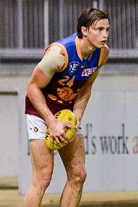 A man wearing a maroon, blue and gold jumper stands on grass.