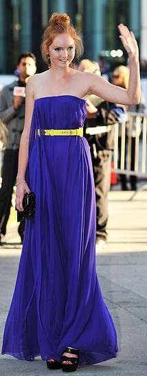 Cole outside wearing a strapless purple dress with her hair up in a large bun, surrounded by photographers