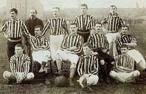 Eleven young men in striped shirts and plain shorts and one older man wearing a suit pose for a team photo. Three sit on the floor, two cross-legged. Four sit on chairs behind, each in a different, casual pose. One has his foot on a football. At the back, the older man and the remaining young men stand, hands on hips or resting on the chairs.