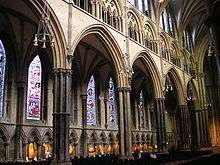 View of the north side of the nave at Lincoln showing wide arcade arches supported on tall slim columns surrounded by shafts of dark marble. The aisle windows are full of stained glass. Beneath them runs a wall arcade in which is displayed a modern set of Stations of the Cross carved in different recycled timbers.