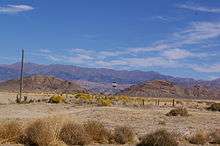Sagebrush and tumbleweed lined desert with two mountain ranges visible in the distance. In the middle of this emptiness is a solitary sign post with a red,white, and blue sign.