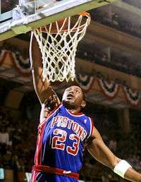 A man, wearing a blue jersey with a word "PISTONS" and the number "23" written in the front, is trying to dunk a basketball.