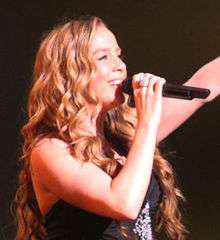 Photograph of Lisa Lavie singing with right hand holding microphone and left arm upraised
