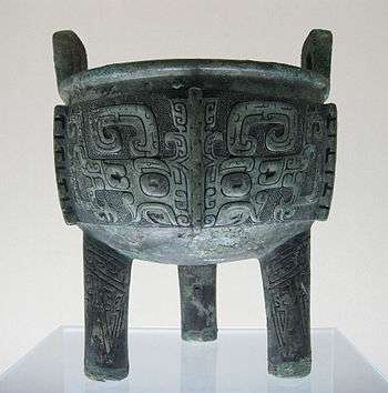 A heavily tarnished bronze bowl adorned with several carvings of squares that curl in on themselves at the bottom. It has three stubby, unadorned legs and two small, square handles coming off from the top rim.