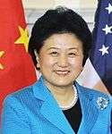 a smiling, aged woman, wearing a blue dress (and a black shirt under it), white necklace and standing in front of the American and Chinese national flags
