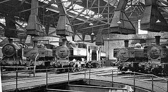 The inside of an engine shed with part of a turntable in the foreground. A number of engines are arranged round the turntable, each with their chimneys under large ventilation cowls.