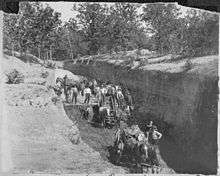 Men and horses hitched to wagons are in a large trench. One wagon is full of coal.