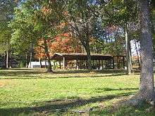 Leaves are turning red on trees in a park graced with a covered picnic shelter on a wide green lawn. Homes are visible on the far side of the park.