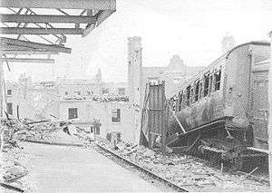 A railway platform strewn with rubble. Broken girders jut out above the platform, while alongside the platform a set of railway lines stop abruptly at a large crater. Next to the railway line is a burned-out railway carriage tilted at an acute angle.