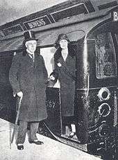 A smartly dressed middle aged gentleman in top hat, velvet-trimmed coat and stripped trousers with spats and rolled umbrella stands next to the open door of the driver's cab of an underground railway train in a station tunnel. A young woman (his daughter) in a long coat and cloche hat stands in the cab doorway.