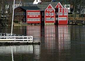 Three bright red buildings stand across the river from the photographer; one of them has a sign saying "Lowell's Boat Shop".