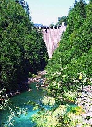 An arch dam straddles a narrow, forested canyon above a tumbling river