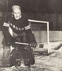 The mask-less ice hockey goalie stands in the crease wearing the small leg pads and gloves of yesteryear.