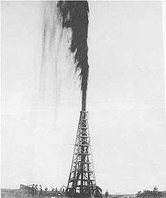 A black-and-white photograph of an oil derrick with a large gusher of oil shooting out the top
