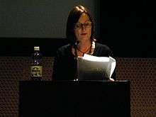 A woman standing on a podium, reading notes from a bunch of papers.