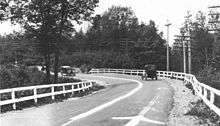 An old photograph of a bend in a road surrounded by trees and power poles. There are wooden guardrails on either side of the road, with a white-painted center line separating the two lanes of traffic. Two old cars are approaching the curve which also has arrows to denote the direction of traffic.