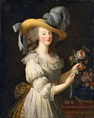 Three-quarter length portrait of a woman wearing a large straw hat with grayish-blue ostrich feathers and a ruffly cream dress with a gold bow tied at the waist in the back. She is arranging a group of pink flowers.