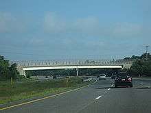 Ground-level view of a busy, six-lane divided freeway; a large overpass bridge is visible in the distance.