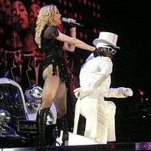 A faraway image of a female blond woman on stage in front of a white Speedster. The woman is wearing a white hat, black top and dancing. Two huge backdrops display two African American males in front of blinking lights. The one in the top backdrop looks towards the camera and smiles and the one in the bottom backdrop is looking down while wearing a white-rimmed sunglass.