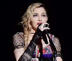 Madonna in a black dress in front of a microphone, holding it with her right hand