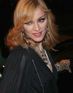 Madonna in a black dress and golden curls, looks towards her right. She wears bangles in her left hand