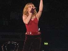 A female blond performer standing on a stage. She is wearing a red top and dark brown pants with a red shiny belt. Her left hand is stretched upwards. She is looking up while holding a microphone on her right hand. Beside her, a chair is visible.