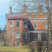 The same house seen from the side with the large wing, also in springtime, illuminated by sun from the left, with some bare branches and bushes partially obscuring the view