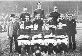 A black-and-white photograph of a football team lining up before a match. Four players, wearing dark shirts, light shorts and dark socks, are seated. Four more players are standing immediately behind them, and three more are standing on a higher level on the back row. Two men in suits are standing either side of the players.