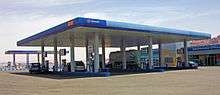 A large gas station with a blue canopy with the words "Manaseer" and the company logo on it. Below it are pumps marked as "diesel" in English and Arabic. In the rear is a large one-story building with "Administration offices" on it