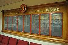 A wooden board headed with the Manchester United crest and the words "International Honours Board", with names in gold writing on eight slate panels, protected by perspex panels.