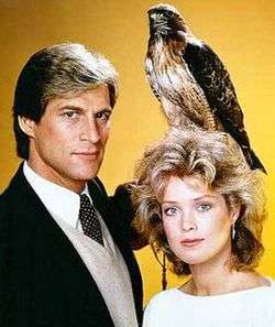 A man in a suit, holding a hawk. Beside him a blonde woman.