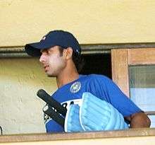 A man in the blue Indian cricket practice kit, wearing a cap carrying his bats and batting pads.
