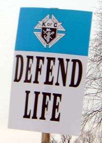 A photograph of a placard at the March of Life that reads defend life on the bottom with the emblem of the Order in a blue band on top.