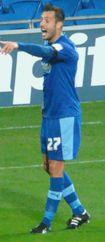 A man wearing blue shirt, shorts and socks, standing on a grass field