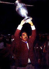 Photo of a man with a moustache, wearing a suit, triumphantly raising a trophy