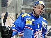 An ice hockey player stands looking to the right of the camera. He is wearing a blue and yellow heltmet and a blue uniform with a large white 7 on his chest.